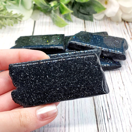 lady's hand holding one small rectangular specular hematite with a stack of more specularite behind her hand